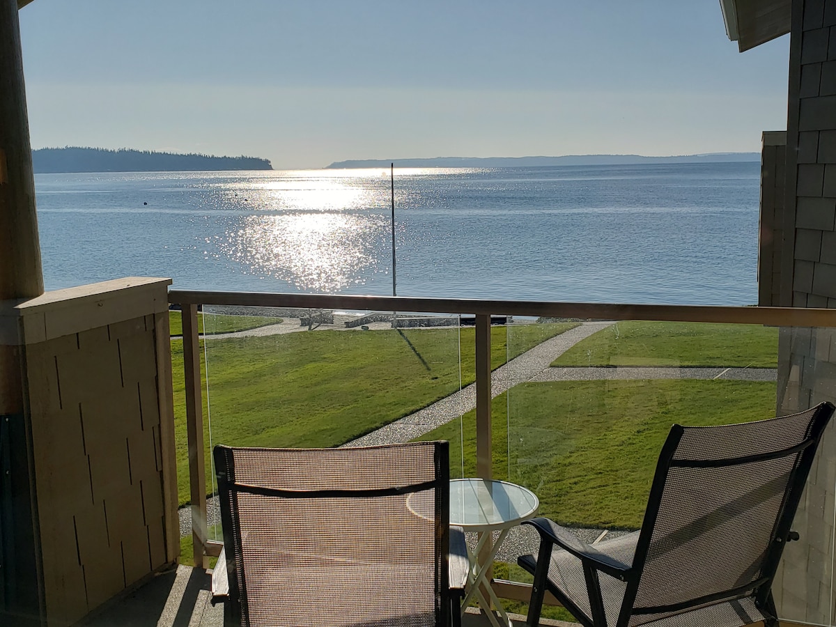 A serene view from a balcony offers glimpses of sparkling water under sunlight, with a grassy area and a stone pathway visible below. Two comfortable chairs face the water, accompanied by a small round table, creating a tranquil outdoor seating arrangement.