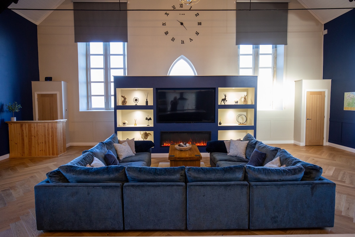The open-plan living area is characterized by a large blue sofa arranged in a U-shape, centered around a wooden coffee table. A wall-mounted TV is flanked by built-in shelves containing various decorative items. The space is illuminated with natural light from the large windows.