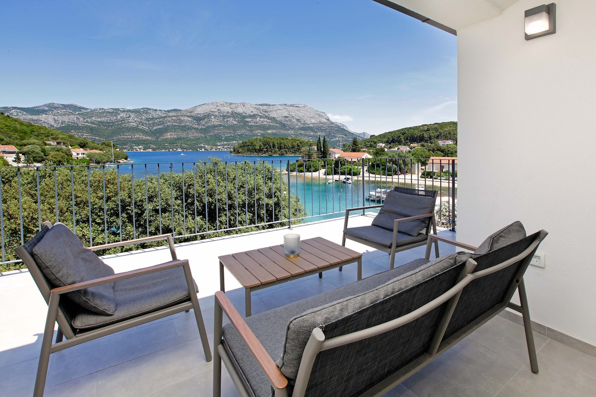 A spacious balcony features two gray lounge chairs and a small wooden table. The scenic view of the bay showcases vibrant blue water and lush greenery, framed by distant mountains under a clear sky.