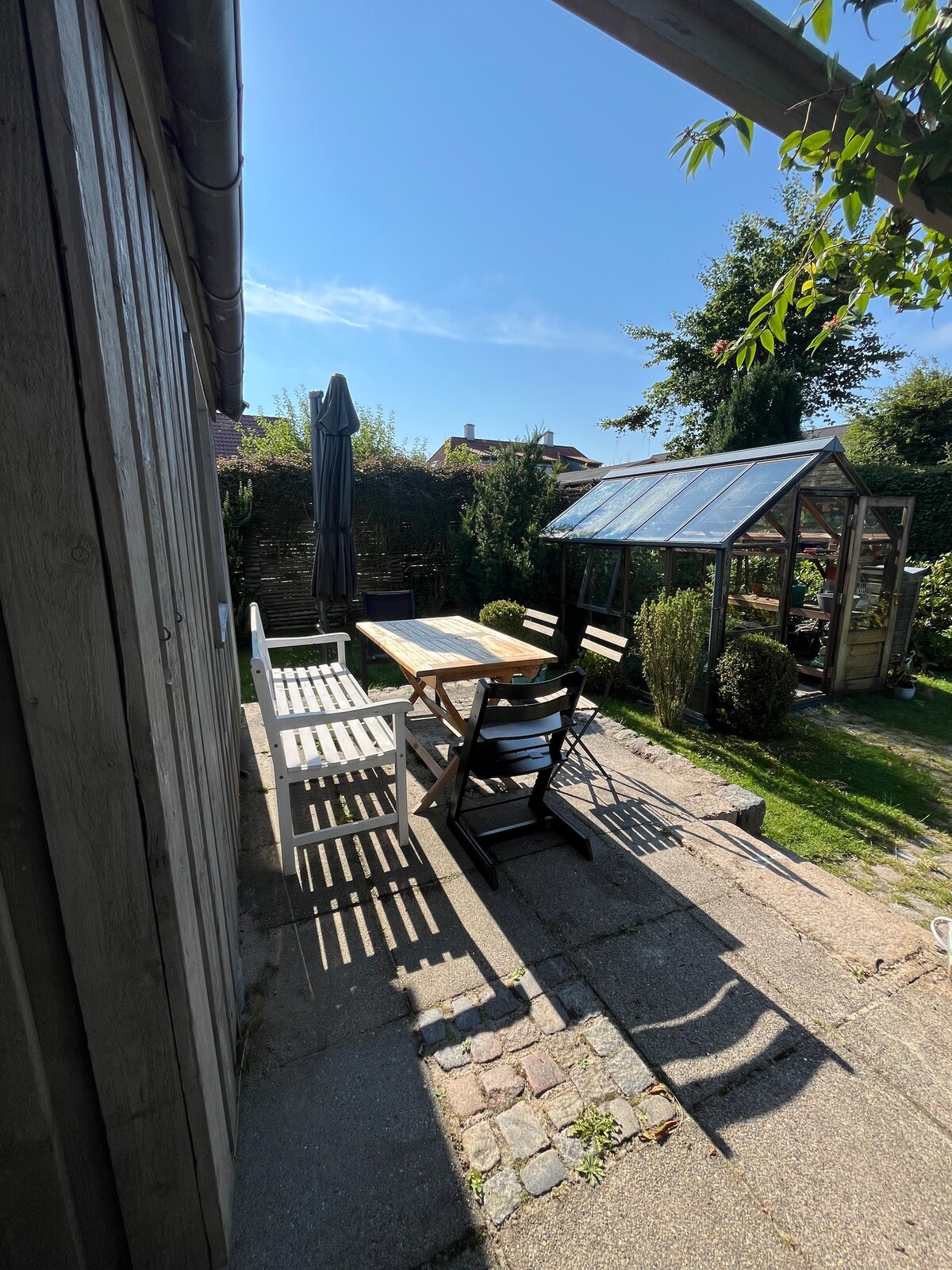 A sunlit patio area features a wooden dining table surrounded by black and white chairs. Shadows from nearby trees create a pattern on the stone paving. A greenhouse is visible in the background, complemented by greenery and a clear blue sky.