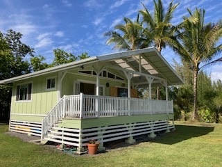 A quaint one-bedroom cabin is showcased with a spacious open deck, surrounded by lush greenery. Tall palm trees stand nearby, complementing the clear blue sky above. The cabin features a light green exterior and a welcoming entrance with stairs leading to the deck.