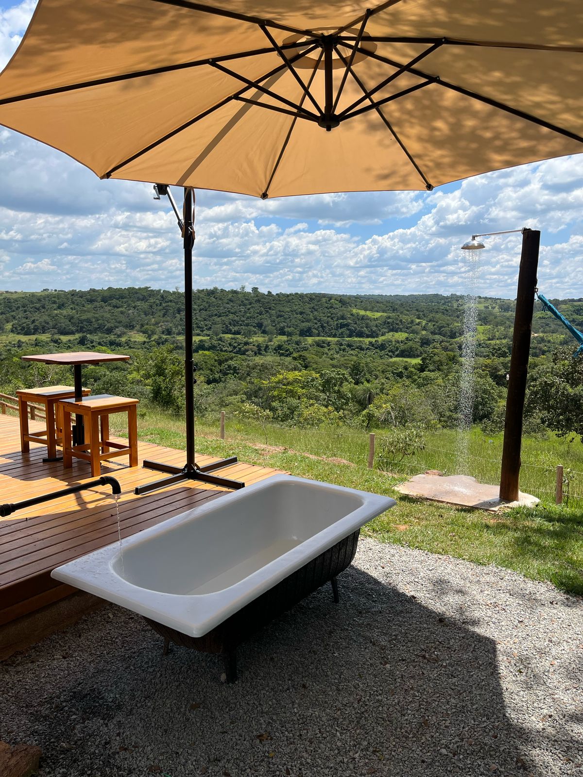 An outdoor relaxation area features a vintage bathtub beneath a large umbrella, offering shade. A wooden deck with two bar stools provides seating. In the background, scenic views of rolling hills and trees are visible, enhancing the tranquil setting.