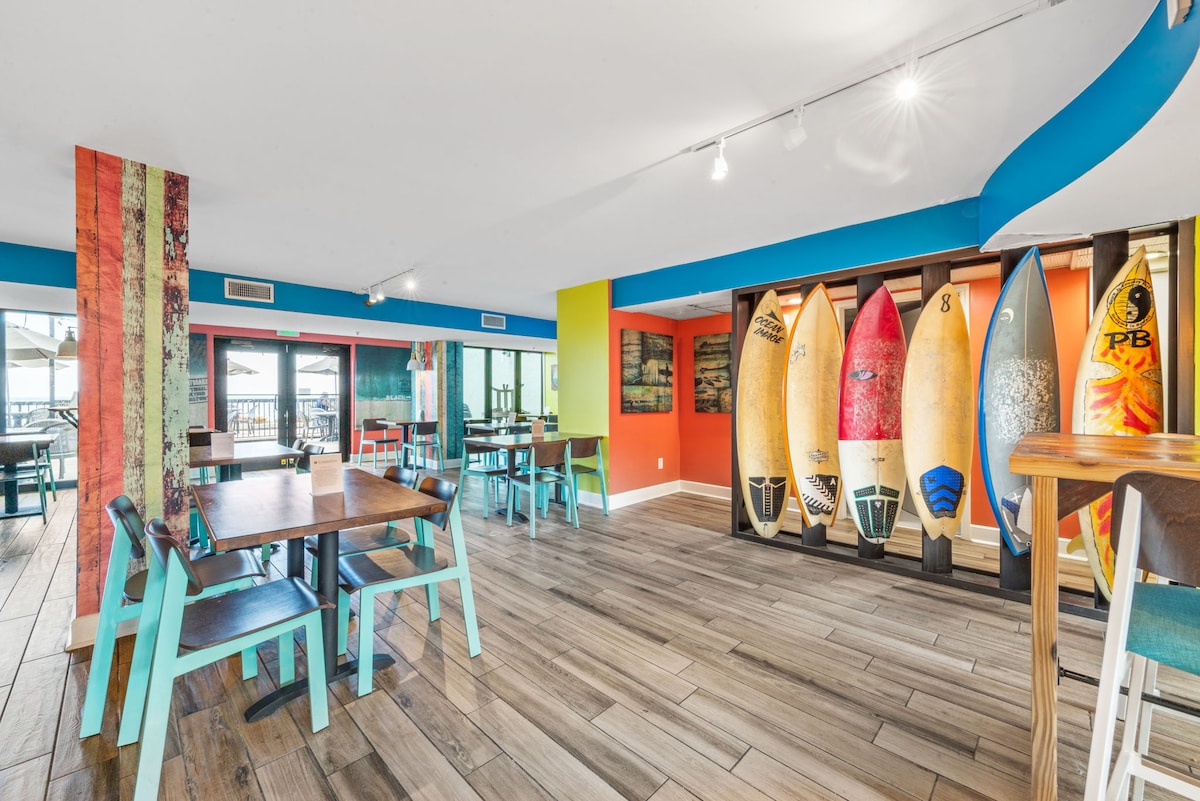 A vibrant dining area features colorful walls with surfboards displayed prominently. Wooden tables and chairs provide seating options, while large windows allow natural light to flood the space, offering views of the outdoor patio.