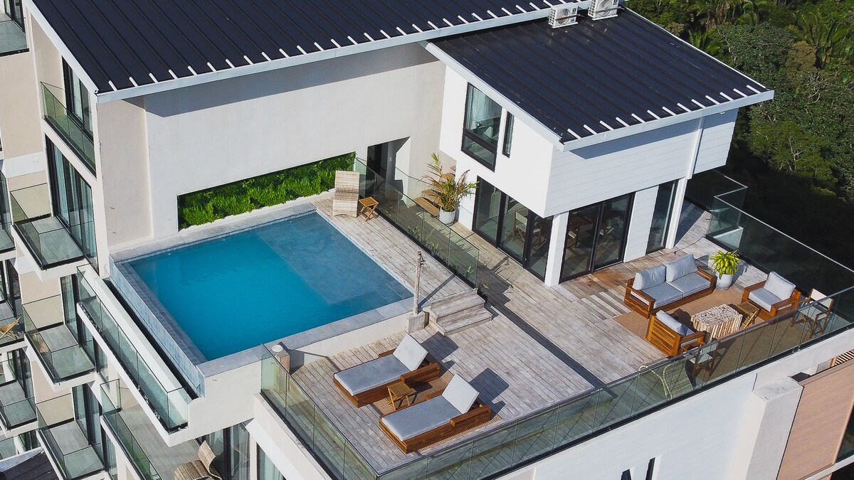 An aerial view of a modern rooftop terrace featuring a private pool, surrounded by wooden lounge chairs and a seating area. Lush greenery is visible in the background, providing a natural contrast to the sleek design of the condo.