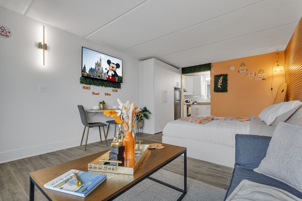 An inviting studio space features a modern layout with a comfortable sofa bed and a spacious king-sized bed. A large TV is mounted on the wall displaying a playful image. A minimalist dining table is positioned beside the kitchenette, enhancing the functional design.