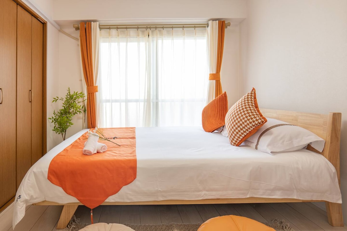 A well-lit bedroom features a spacious bed adorned with crisp white linens and an orange bed cover. Decorative pillows add a pop of color. Natural light flows through large windows with sheer curtains, while a small plant sits on the bedside, enhancing the serene environment.