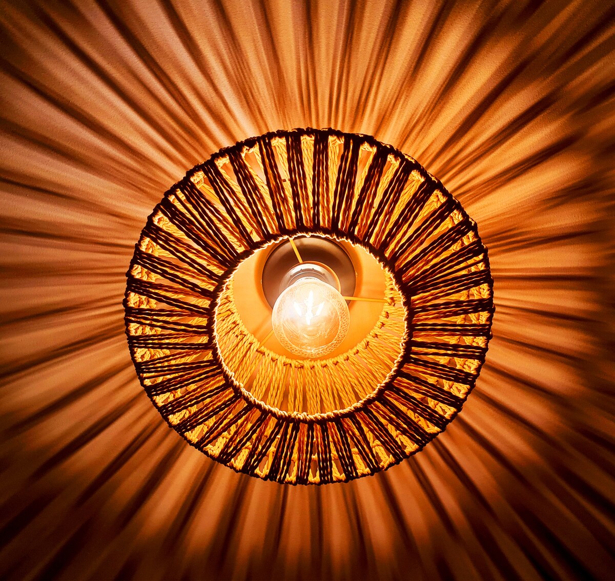 A circular light fixture is captured from below, featuring a woven design that radiates downward. The warm glow from the exposed bulb creates a soft ambiance, with light rays spreading in a circular pattern across the ceiling.