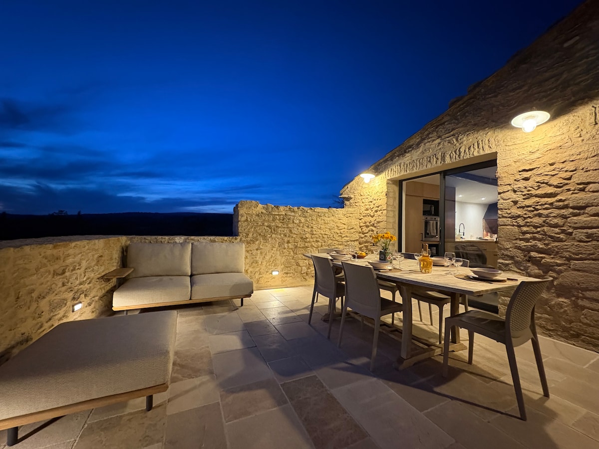An outdoor terrace is presented under a darkening sky, featuring a dining table surrounded by several chairs. A comfortable lounge area is visible, complemented by soft cushions. Warm lighting enhances the stone walls, while a serene ambiance is created by the open view of the landscape.