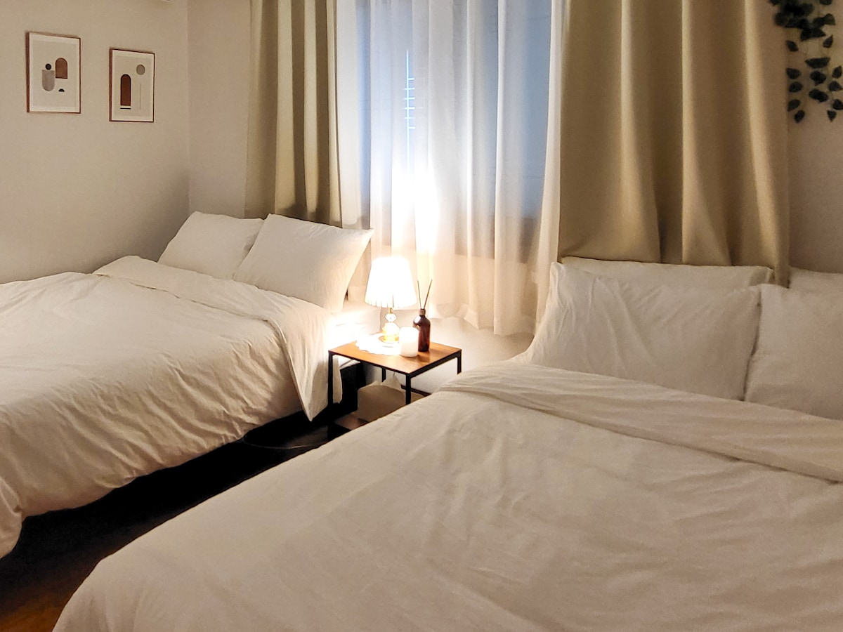 The cozy bedroom features two spacious beds dressed in crisp white linens. A soft lamp provides gentle lighting beside a small bedside table. Sheer curtains gently diffuse natural light, creating a serene ambiance in the room.