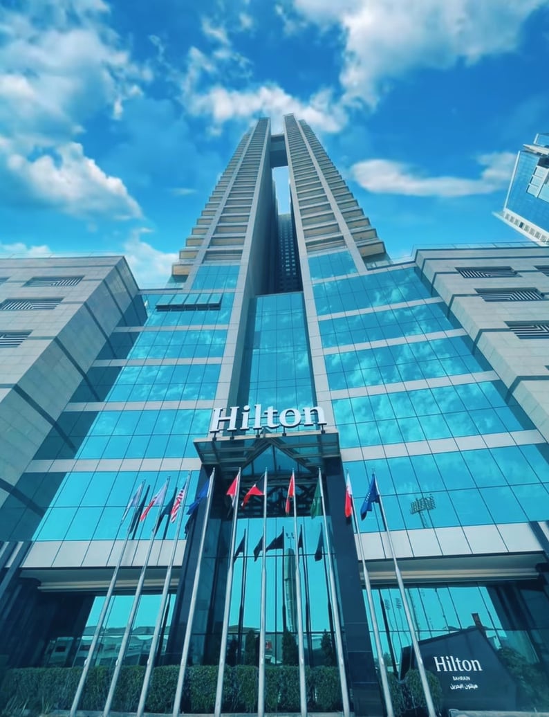 The modern building features a sleek glass façade reflecting the sky. Prominent Hilton branding is displayed at the entrance, accompanied by multiple flags. The architecture includes two tall towers, creating a striking vertical presence against a backdrop of scattered clouds.