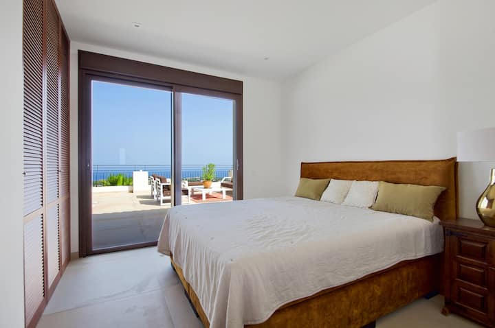 Priestranná, elegantná spálňa s úchvatným výhľadom na more cez sklenené dvere od podlahy až po strop, ktoré sa otvárajú na veľkú terasu. Izba je kúpaná v prirodzenom svetle a vytvára pokojnú a vzdušnú atmosféru.