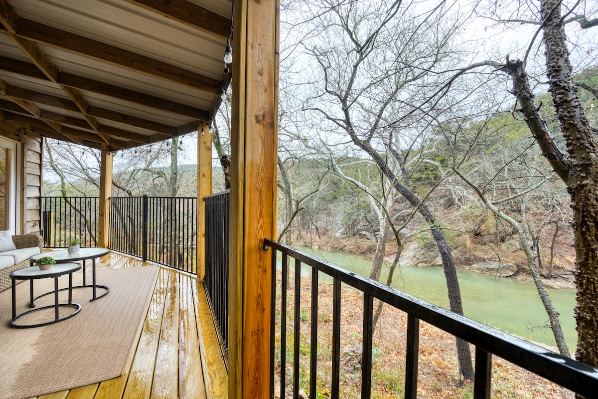 The covered back deck features wooden flooring and a metal railing, providing a clear view of the flowing creek below. Two modern tables are positioned on a woven rug, while trees with bare branches frame the landscape, emphasizing the natural surroundings.