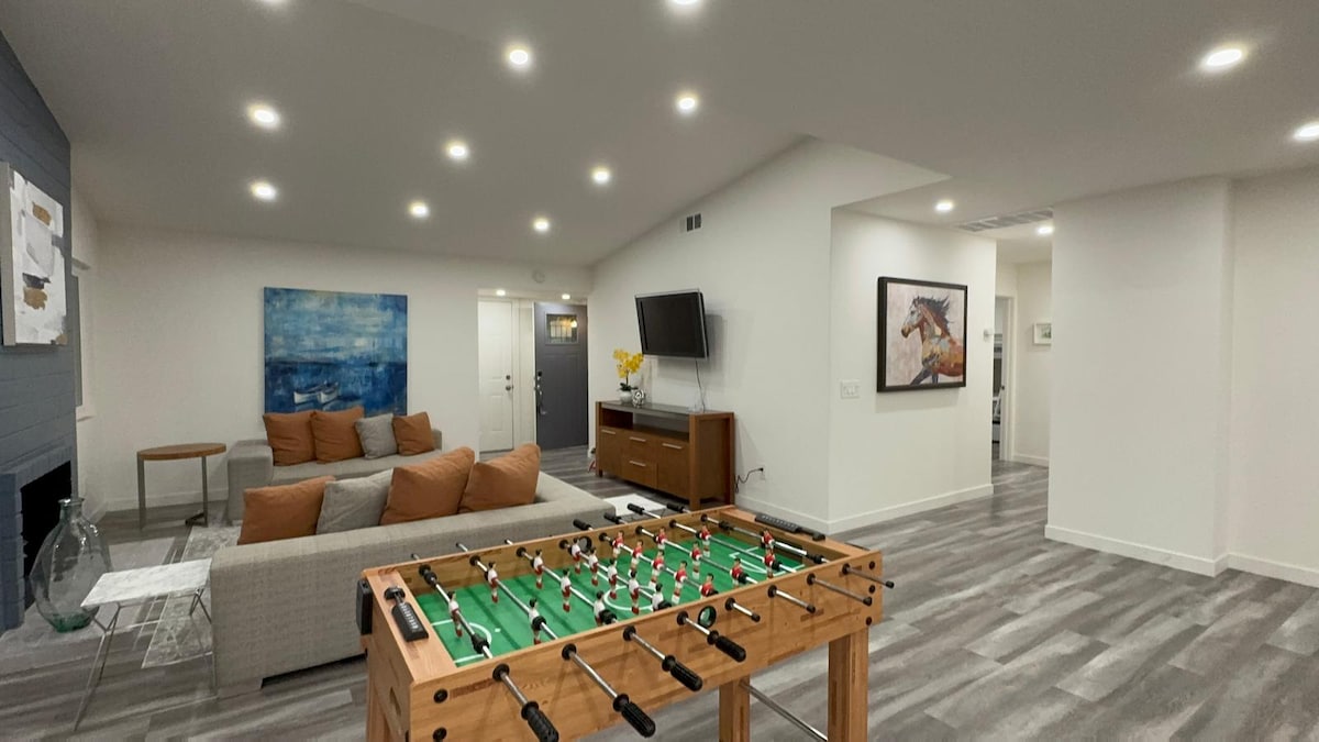 An open game area features a foosball table in the foreground. Soft lighting enhances the spacious living room, where a sectional sofa with cushions and a modern entertainment unit are visible. Artwork adorns the walls, adding interest to the neutral decor.