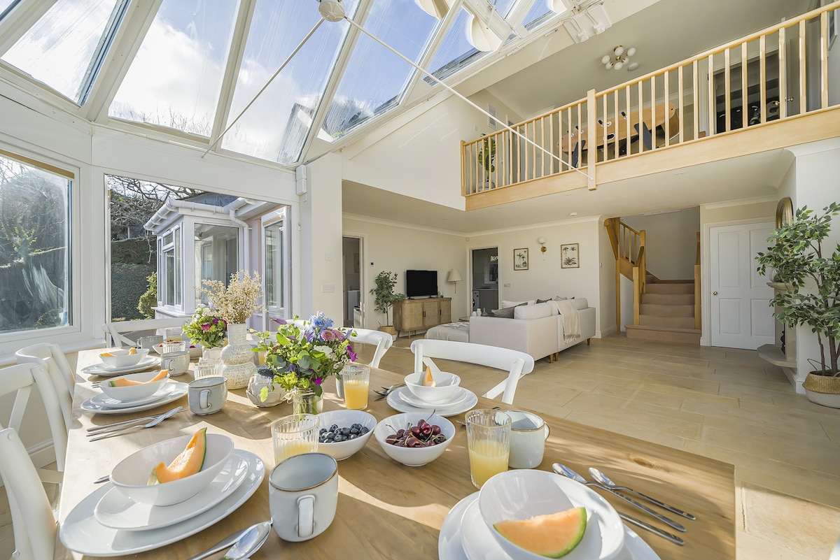 An inviting dining area is shown with a spacious wooden table set for breakfast. Plates feature fresh fruit, and beverages are arranged nearby. A large glass ceiling allows natural light to fill the room, highlighting the open design connecting to the living space and balcony above.