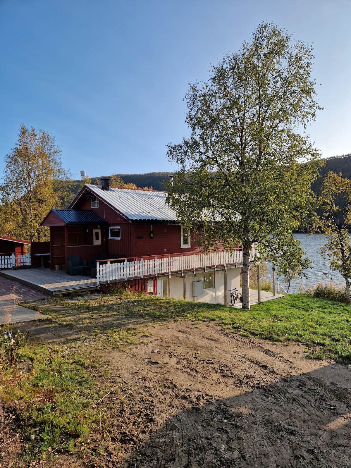 The exterior of a red cabin is shown, featuring a sloped roof and a white railing along a spacious deck. Nearby, a tree provides shade, while the surrounding landscape includes grass and a glimpse of water in the background, set against the backdrop of a forested hillside.