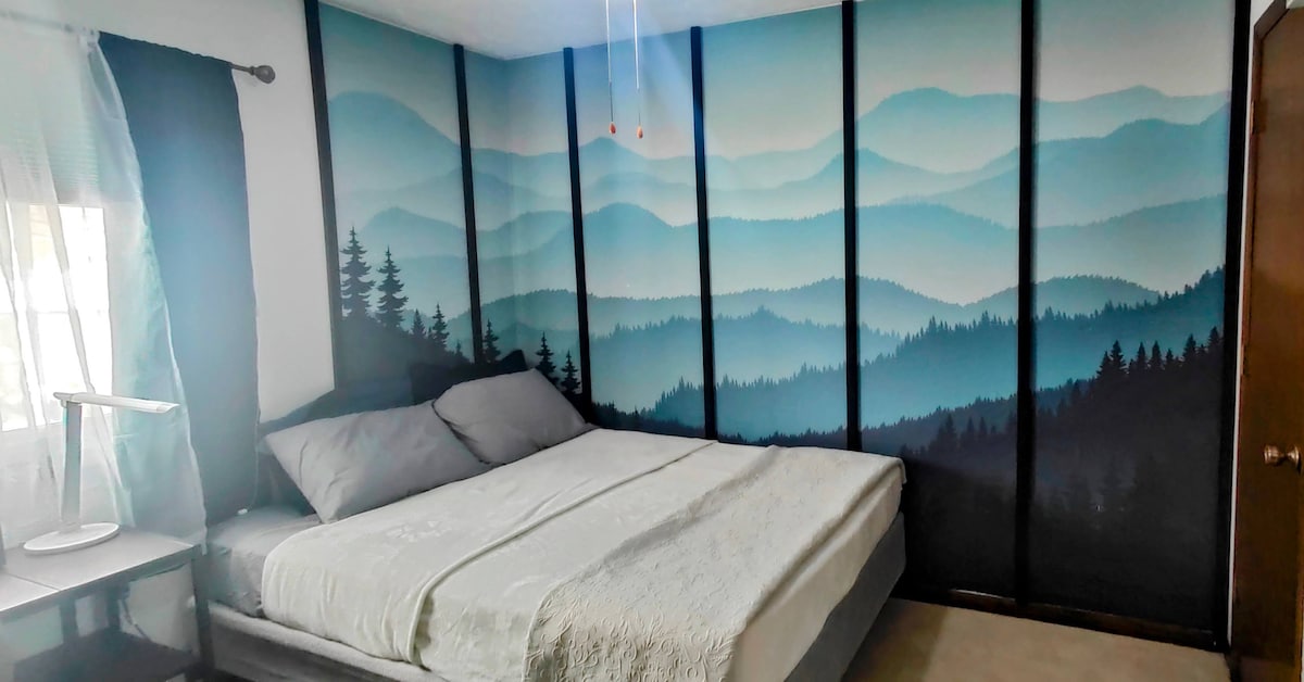 A calming bedroom features a large bed topped with soft gray bedding. A mountain-themed mural in shades of blue covers the wall, complementing the serene ambiance. Natural light enters through a window with sheer curtains, creating a tranquil setting.