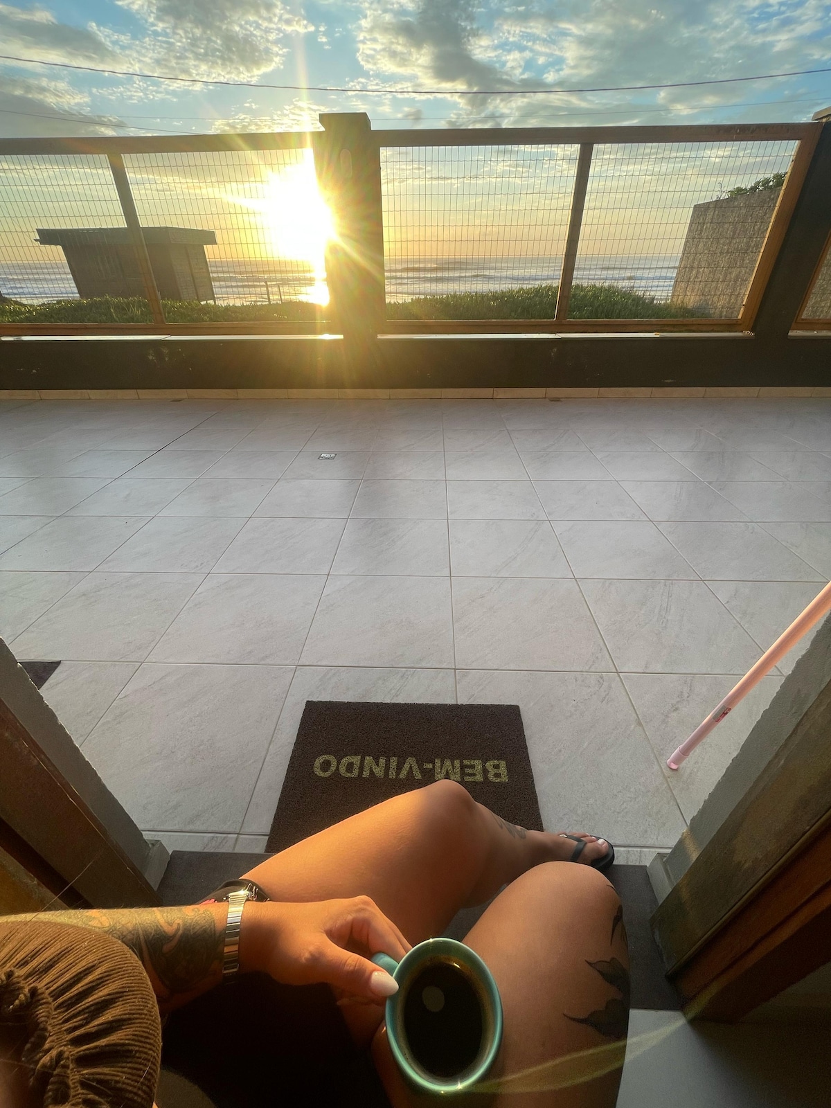 A calming view from a doorway reveals a tranquil sunrise over the ocean. The outdoor space features tiled flooring and a wooden railing. A mug is held gently, suggesting a moment of relaxation while enjoying the scenery.