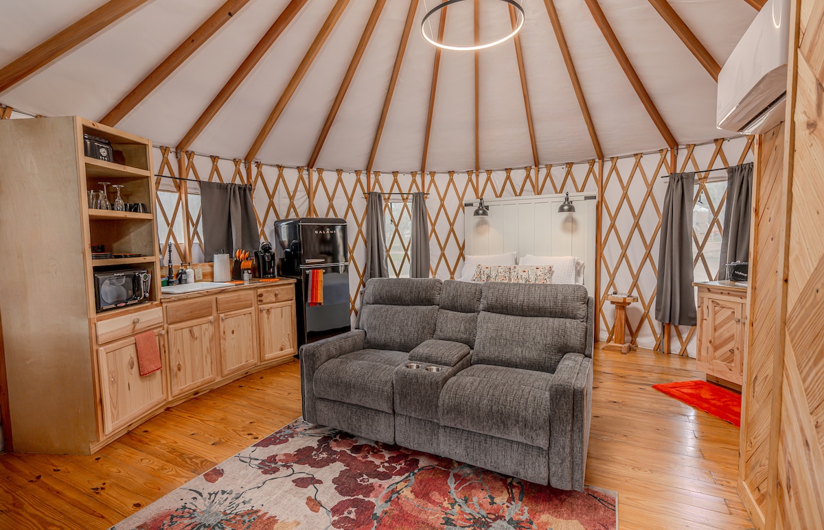 An inviting interior of the Yurt features a comfortable gray double recliner sofa positioned on a patterned area rug. Wooden cabinetry houses a microwave and retro refrigerator, blending rustic charm with modern design. Natural light flows through large windows, enhancing the cozy atmosphere.