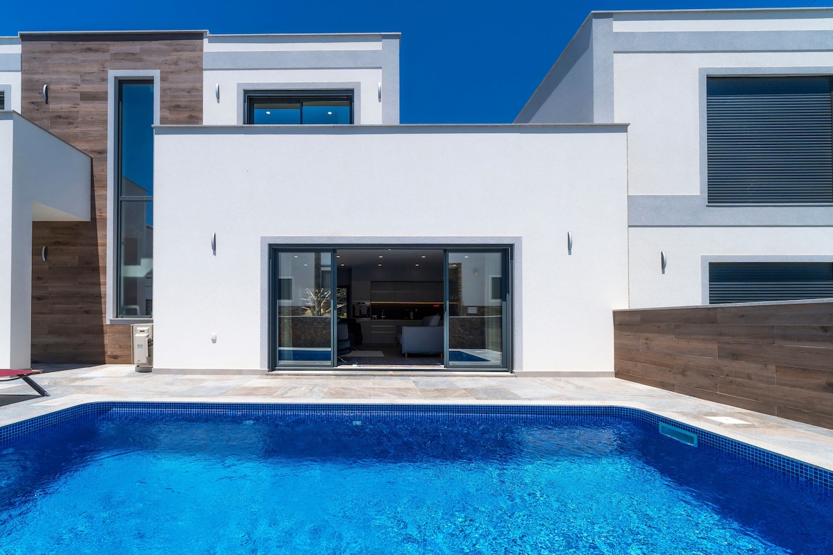 A private pool features clear blue water, surrounded by a spacious patio area. Large glass doors provide a view of the modern interior beyond. The villa's exterior is characterized by a clean, minimalist design and a blend of white and natural wood elements.