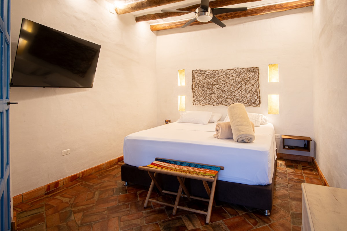A cozy room features a spacious bed with neatly arranged towels. Soft lighting is provided by two wall sconces, while a woven art piece hangs above. A flat-screen TV is mounted on the wall, and a small side table is positioned nearby.