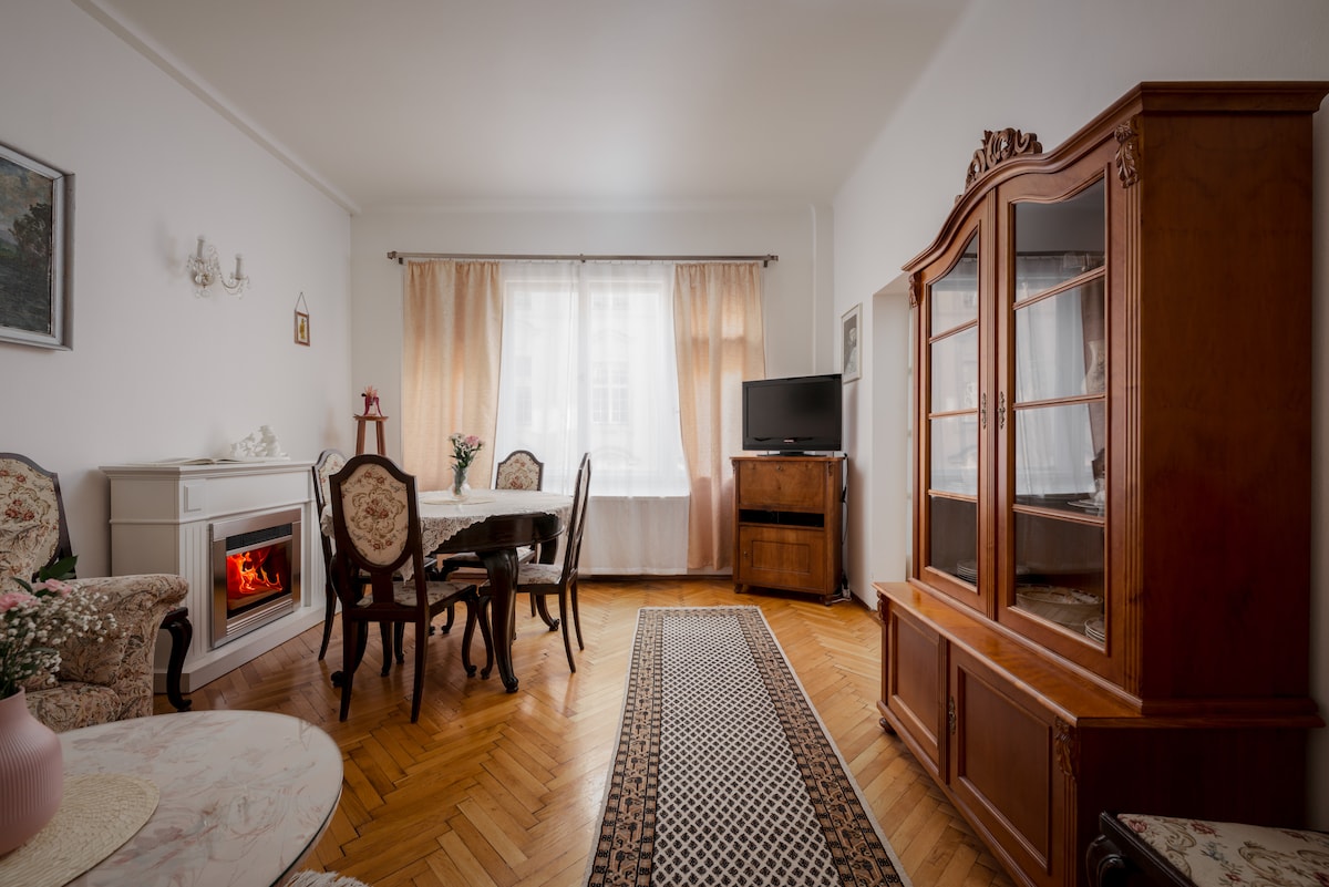 A spacious living and dining area is highlighted by a neutral color palette. A dining table is surrounded by elegant chairs, while a vintage cabinet enhances the traditional decor. A cozy electric fireplace adds warmth, with natural light filtering through large curtains.