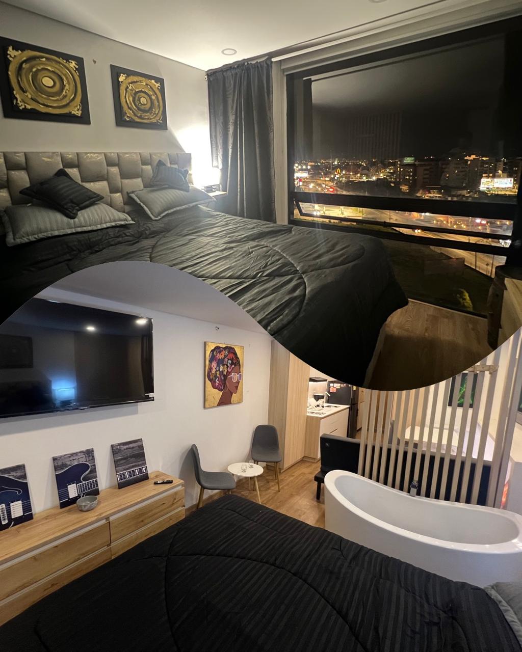 A spacious bedroom features a plush bed with dark bedding, complemented by elegant wall art. Large windows provide a nighttime view of the city lights, while a cozy seating area includes a small table and contemporary chairs, creating a relaxing atmosphere.