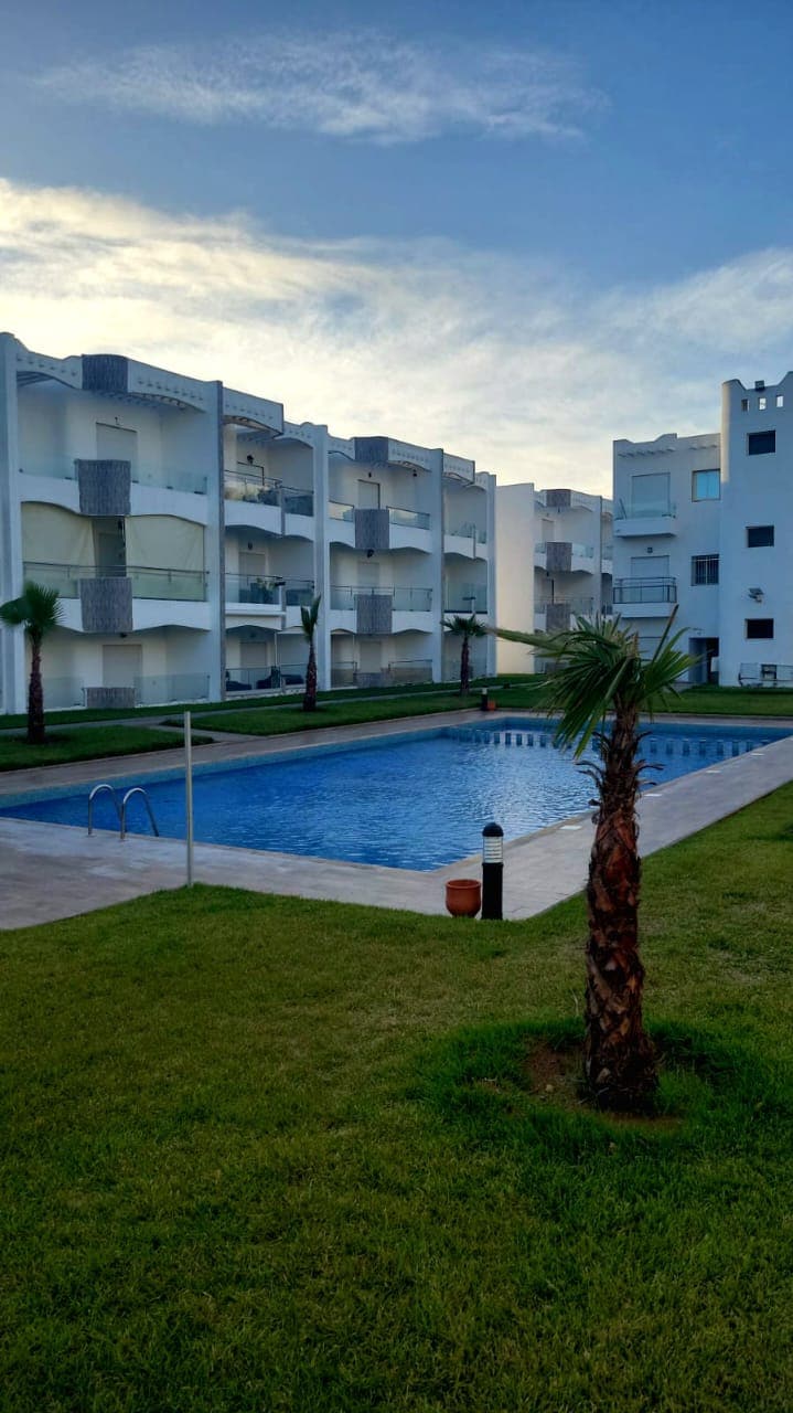 The image displays a serene pool area surrounded by low-rise buildings. A well-maintained lawn and palm trees enhance the inviting outdoor space. The pool reflects the sky, providing a focal point for relaxation. Various balconies overlook the area, ensuring a quiet atmosphere for guests.
