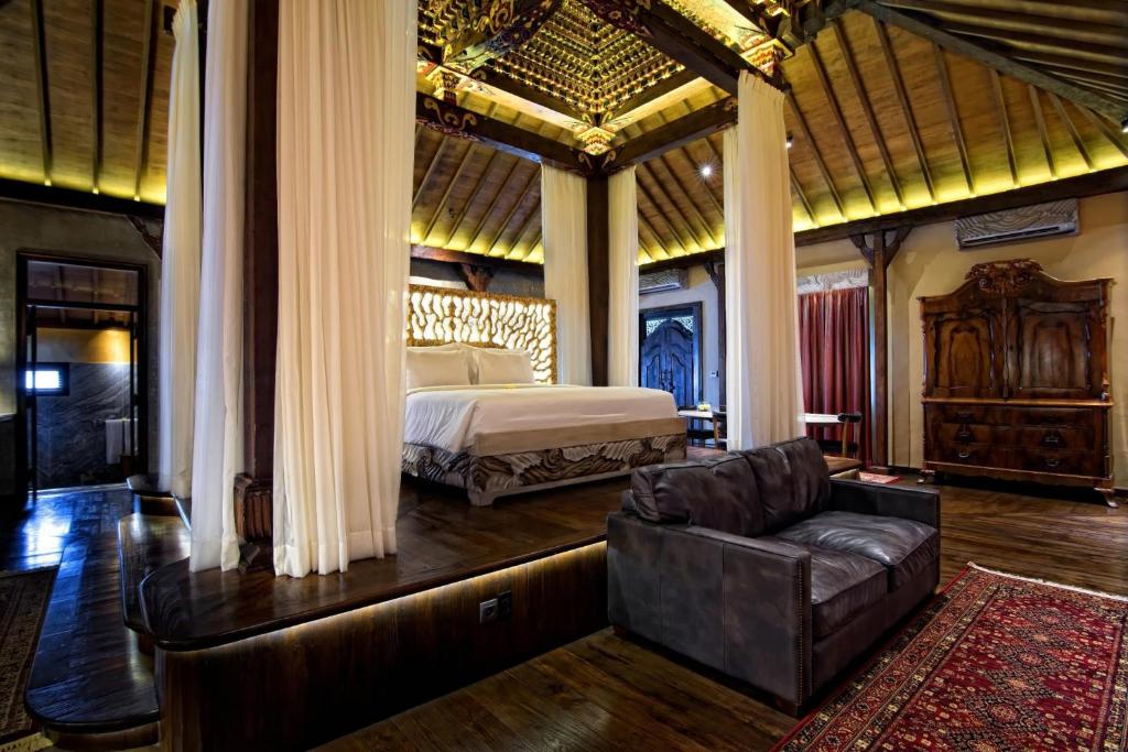 A spacious bedroom features a large elevated bed surrounded by delicate curtains, providing an intimate setting. Soft lighting highlights wooden beams and intricate ceiling details. A comfortable sofa sits on a richly patterned rug, enhancing the room's unique character and inviting relaxation.