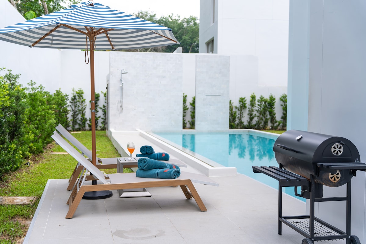A private pool area features two loungers positioned under a striped umbrella, accompanied by rolled towels and a glass of orange juice. A modern grill sits nearby, with a refreshing outdoor shower visible in the background, surrounded by neatly trimmed greenery.