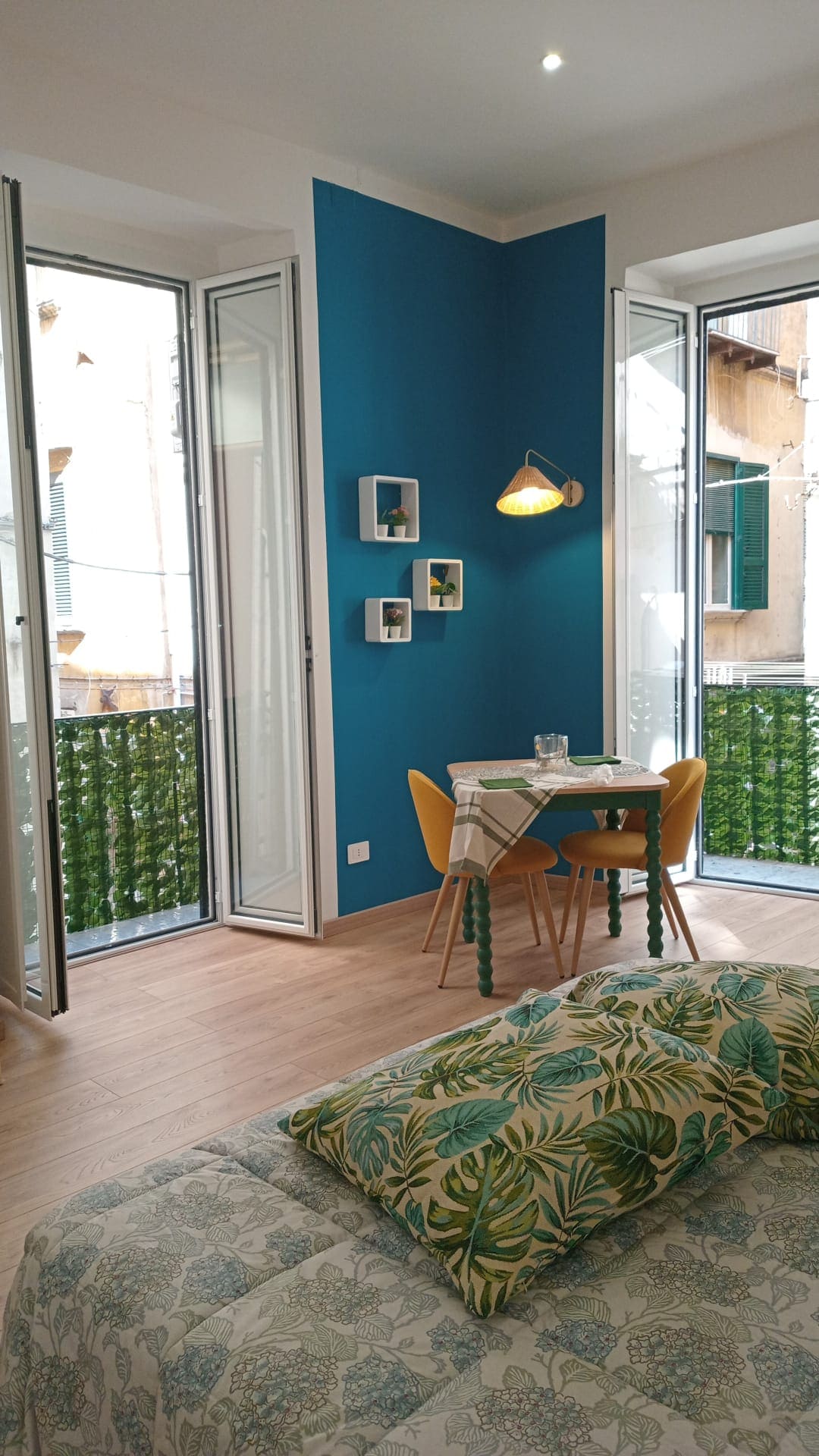 A spacious room is highlighted by vibrant blue walls and large windows offering natural light. A small dining area with a table and yellow chairs is positioned near the window. Decorative wall shelves feature minimalistic items, while a patterned bedspread adds a touch of color.