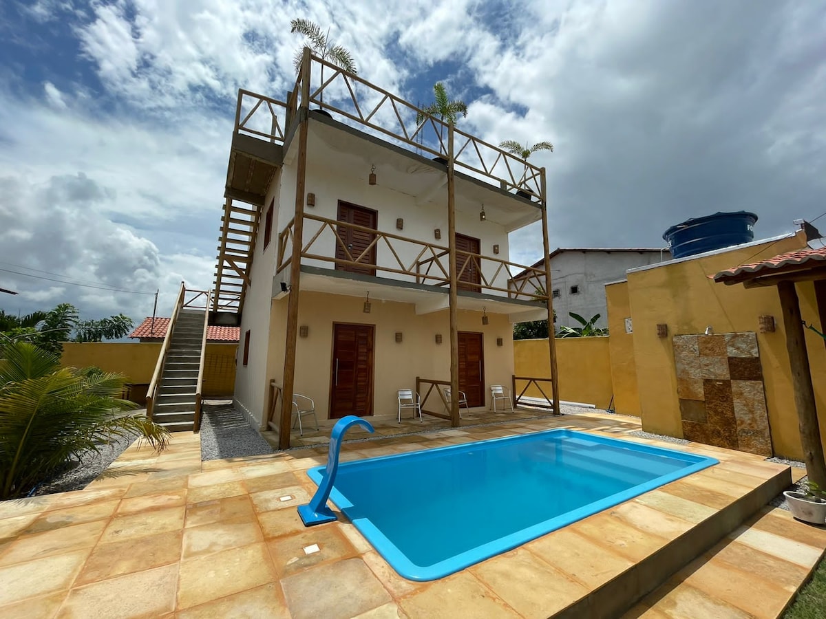 A three-story building is depicted with a wooden railing balcony and exterior stairs leading to the upper floor. A rectangular pool with a light blue interior is centered in the foreground, surrounded by tiled flooring and landscaping elements, including palm plants and a privacy fence.