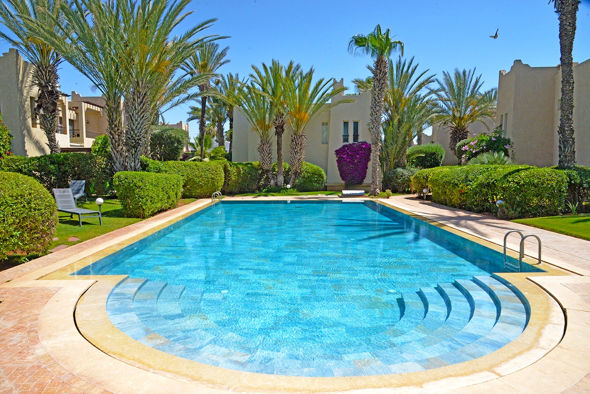 A communal swimming pool is surrounded by well-maintained gardens, featuring tall palm trees and greenery. The pool's clear blue water reflects sunlight, while a gently sloping entry leads to a depth suitable for various activities. Sun loungers are positioned on the pool deck.