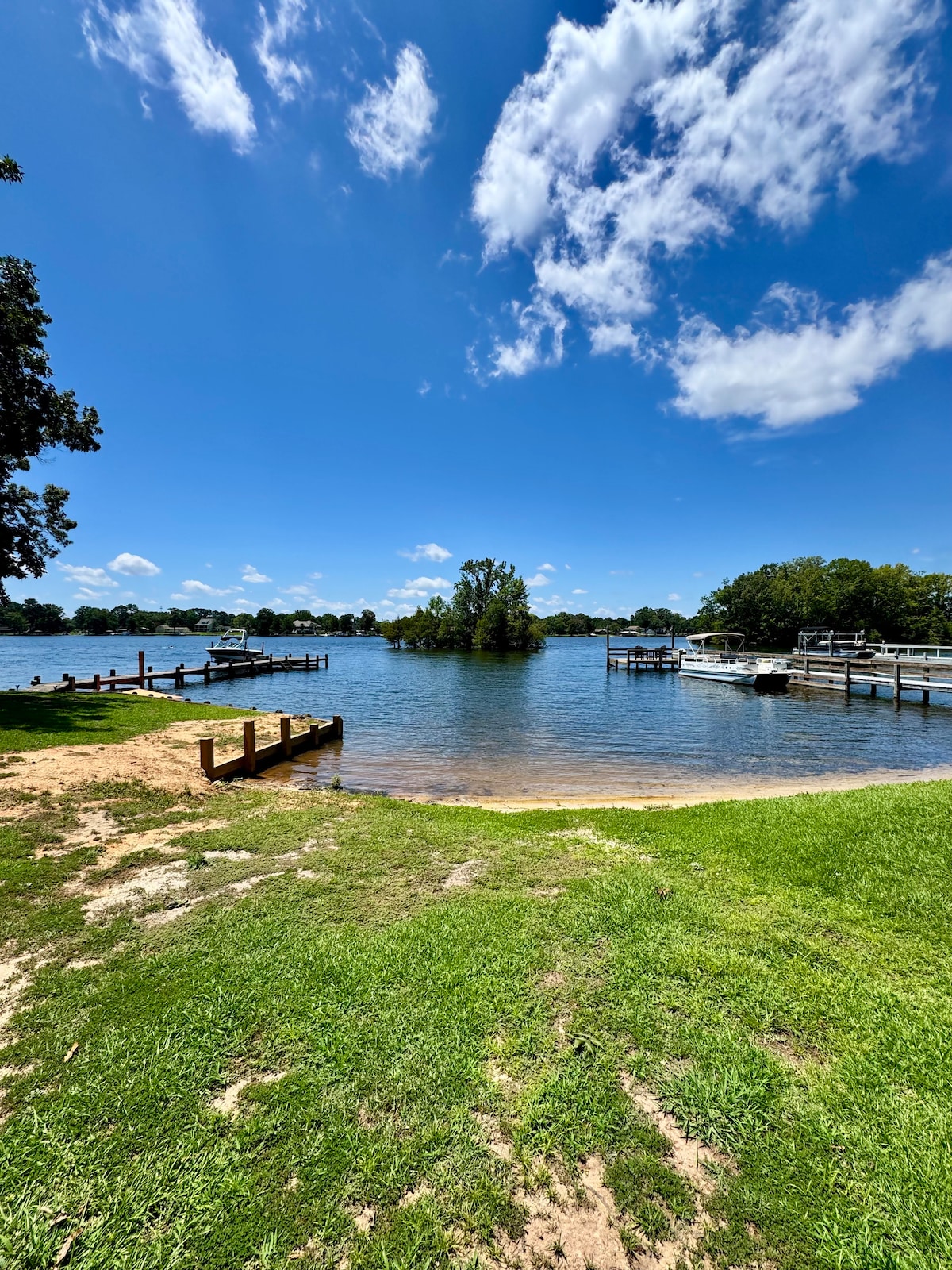A serene lake scene showcases clear blue skies and fluffy white clouds above tranquil waters. Nearby, a grassy area leads to a gentle sandy shore, offering easy access to the lake. Wooden docks extend into the water, with lush greenery in the distance.