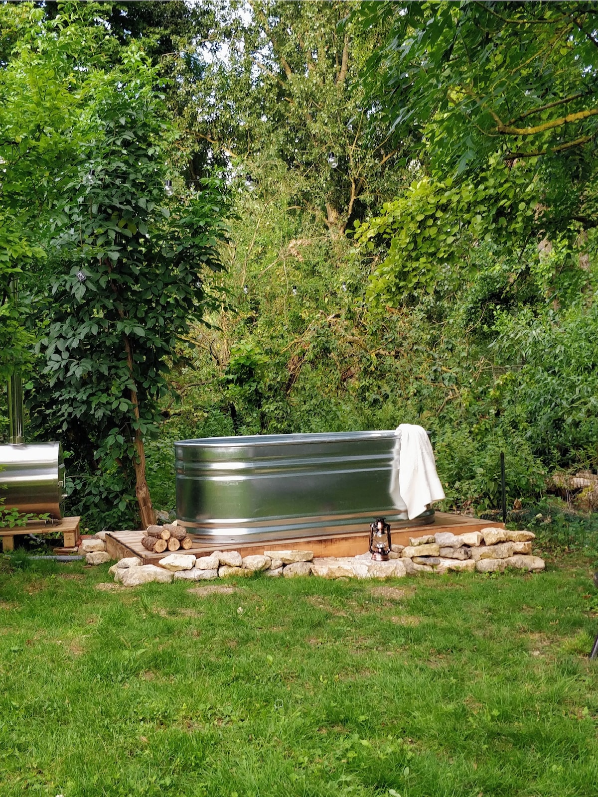 An outdoor wooden hot tub is placed on a stone platform, surrounded by lush greenery. A soft white towel is neatly draped over the edge of the tub. Nearby, a lantern rests on the ground, complementing the serene natural setting.