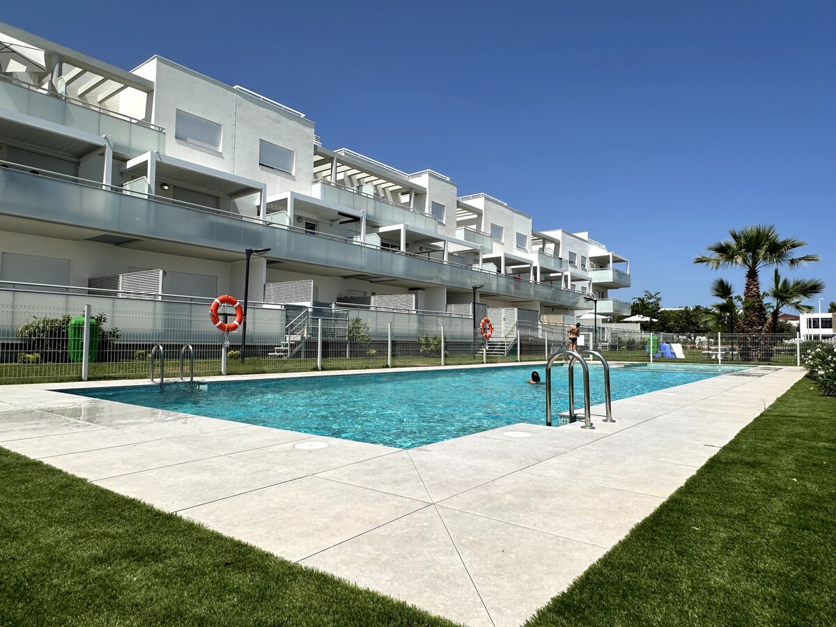 A clear outdoor pool is surrounded by well-maintained grass and features steps for entry. The pool area is enclosed with a fence, and several palm trees lend a touch of greenery. Apartment balconies overlook the pool, contributing to a communal atmosphere.