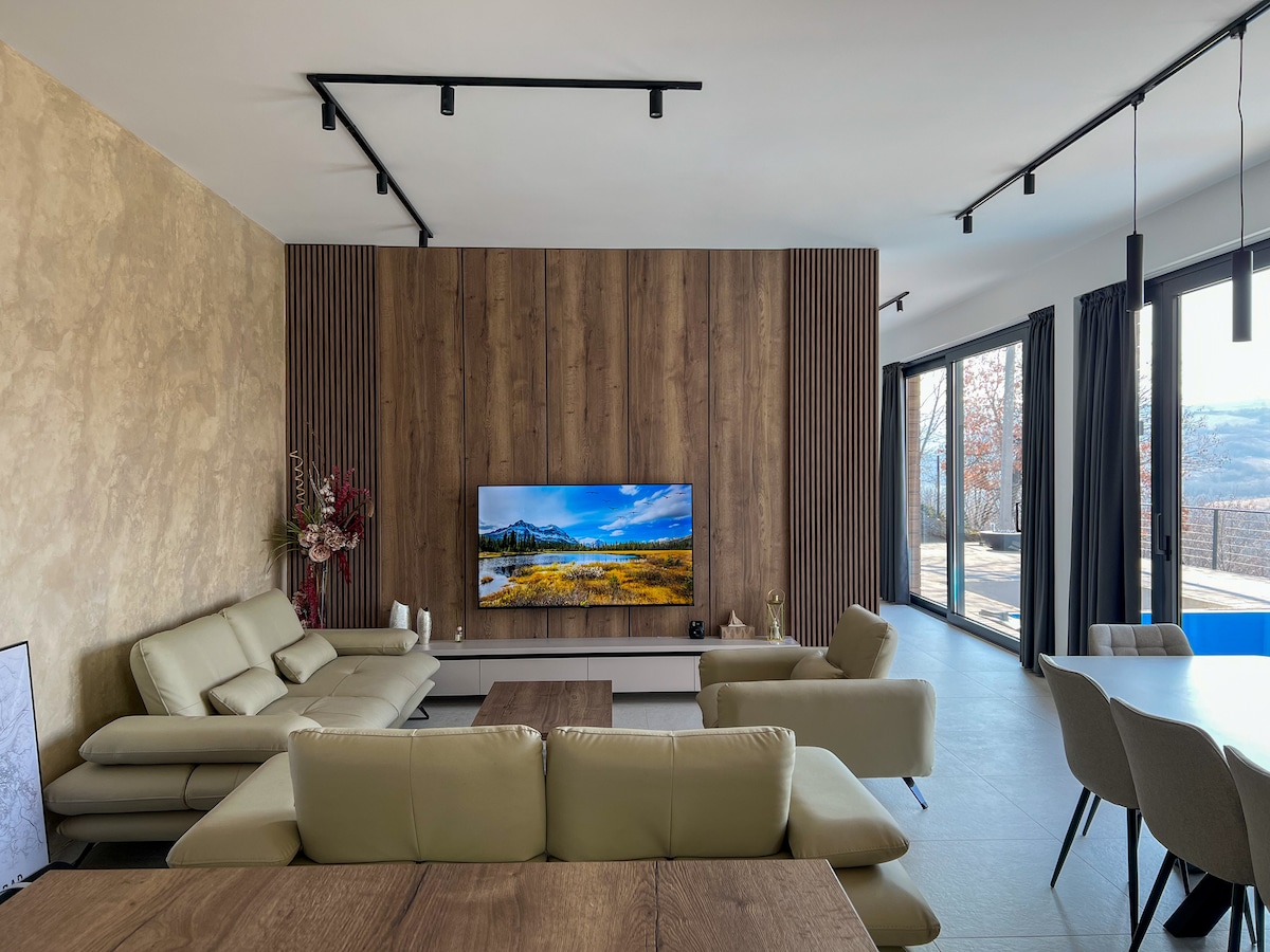 A modern living room features a large flat-screen TV mounted on a wooden accent wall. Light-colored sofas are arranged around a wooden coffee table. Floor-to-ceiling windows provide ample natural light and offer views of the outdoor scenery.