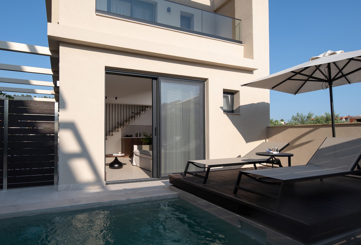 An outdoor area features a private pool beside stylish lounge chairs. A sliding glass door provides access to the interior, while an umbrella offers shade. The building's modern architecture is complemented by a sleek design and green landscape in the background.