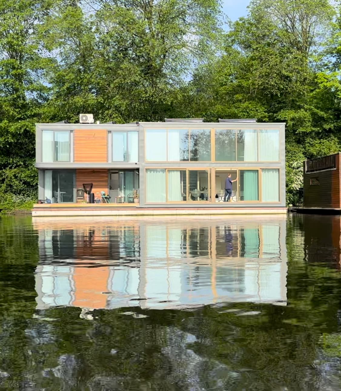 A modern houseboat is positioned on the water's edge, surrounded by lush greenery. Large glass windows reflect in the calm water, allowing natural light to fill the interior spaces. A small outdoor seating area is visible on the terrace, creating an inviting outdoor retreat.
