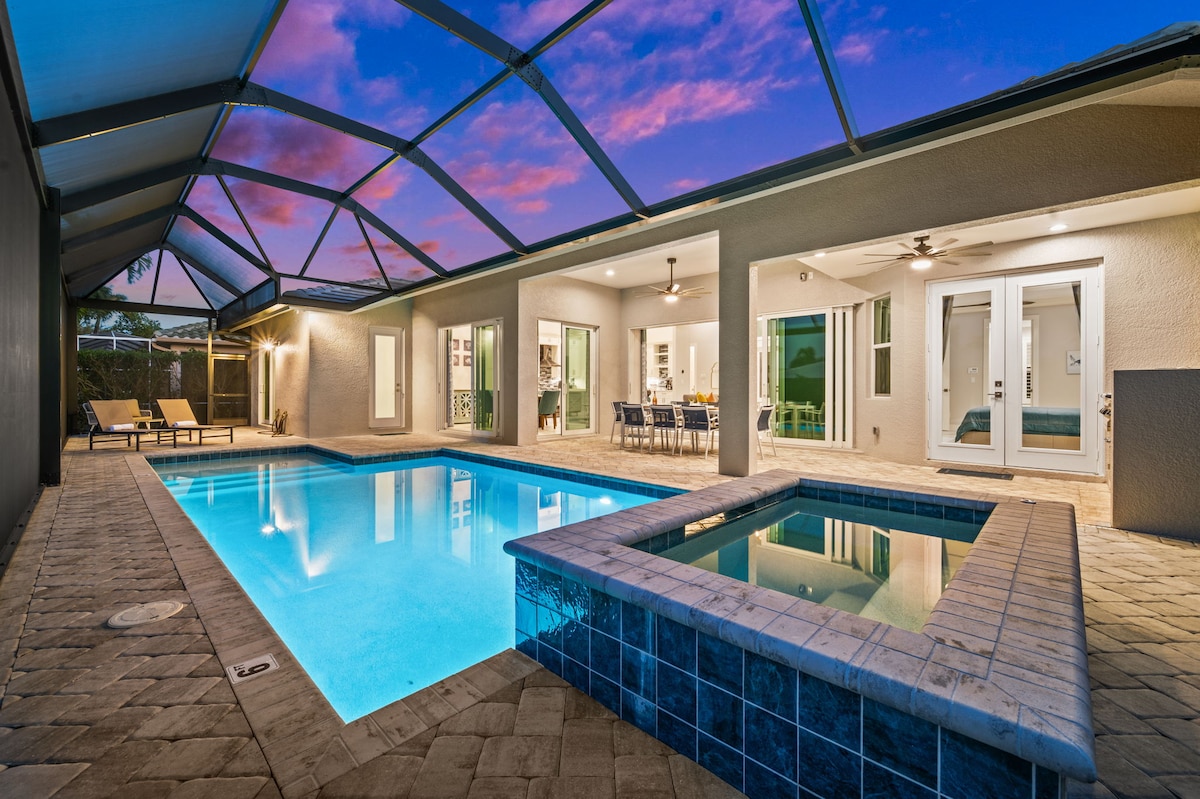 A spacious outdoor area is depicted with a clear blue heated pool and adjoining spa, surrounded by a paved deck. The covered lanai offers seating areas and dining space, with large sliding glass doors leading into the interior living space, framed by a vibrant sunset sky.