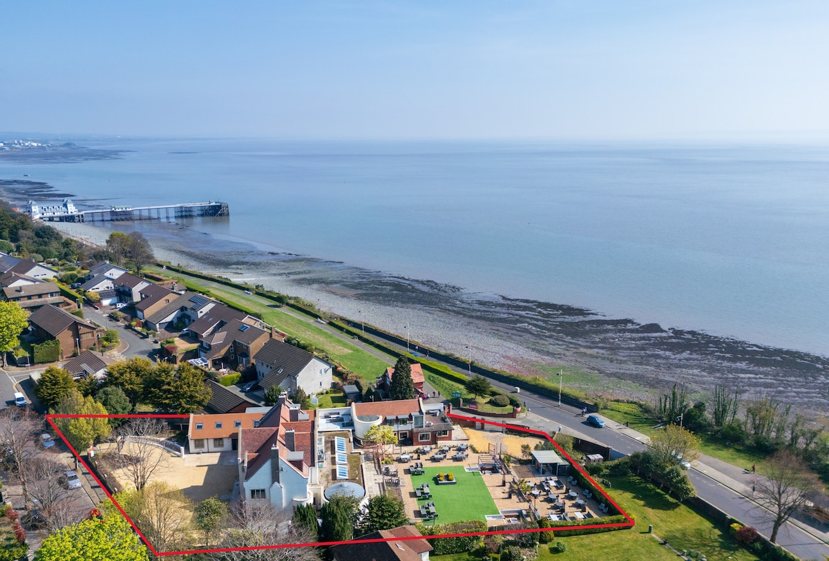 An aerial view captures the coastal property bordered by lush greenery, showcasing well-maintained gardens and seating areas. The Bristol Channel stretches peacefully beyond, with the iconic Penarth Pier visible in the background, illustrating the property's prime seaside location.