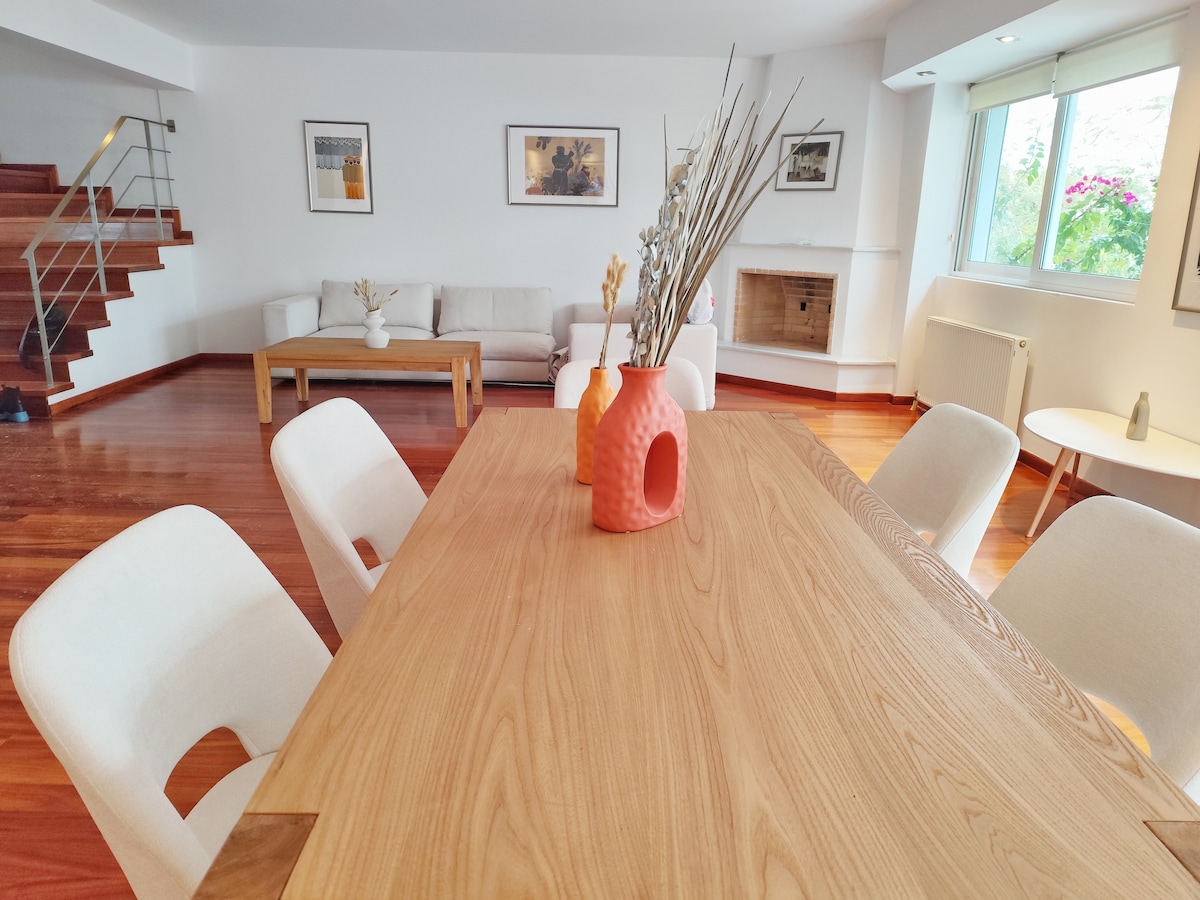 A wooden dining table, set for eight, is positioned in an open space. Soft beige chairs surround the table, while a decorative vase with dried flowers adds a touch of warmth. A comfortable sofa and a fireplace are visible in the background, alongside abundant natural light from large windows.