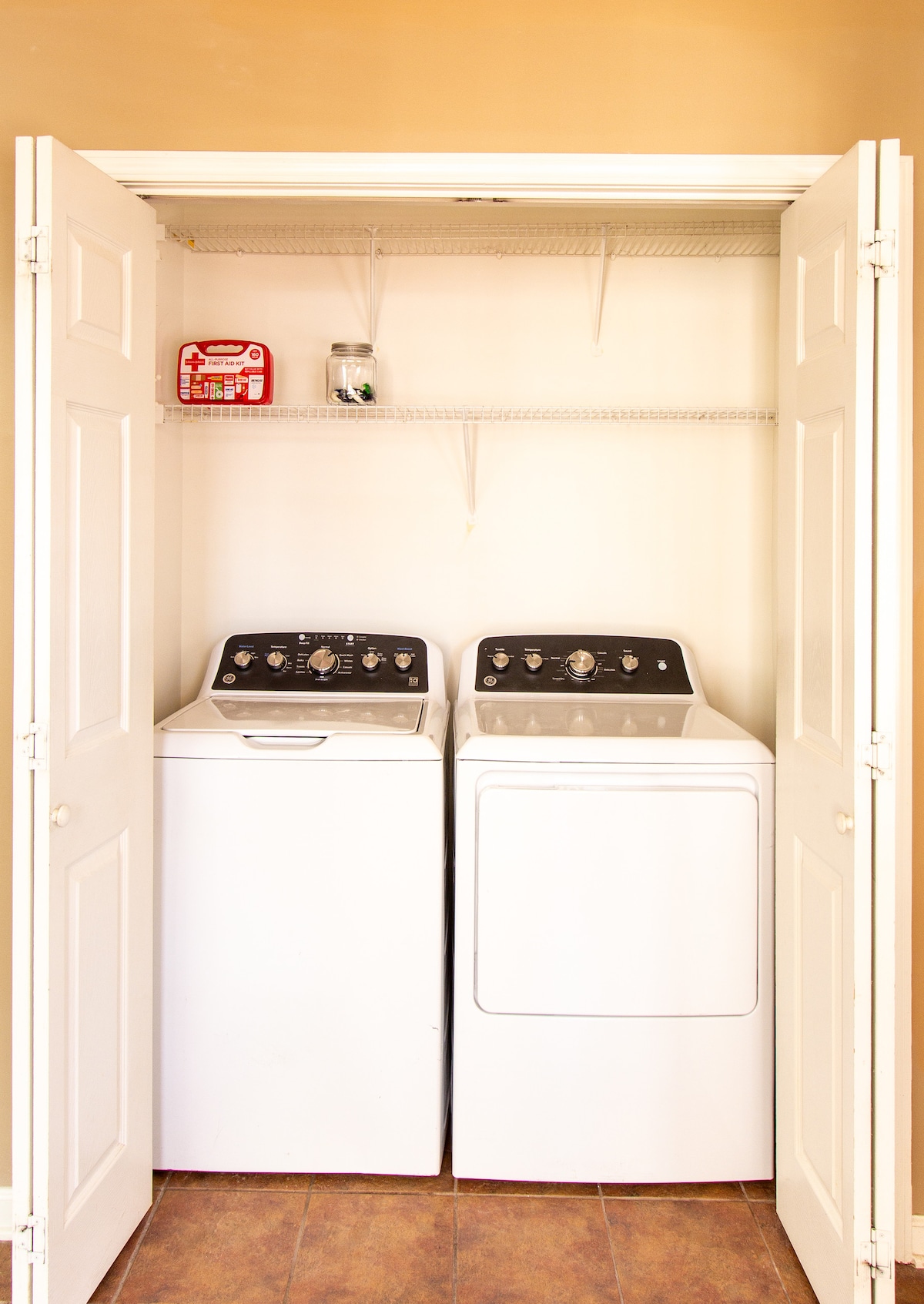 A laundry area is presented with a stacked washer and dryer. Above, a shelf holds a small container and other items. The space is lit with neutral wall colors and a tile floor, providing practicality within the home's layout.