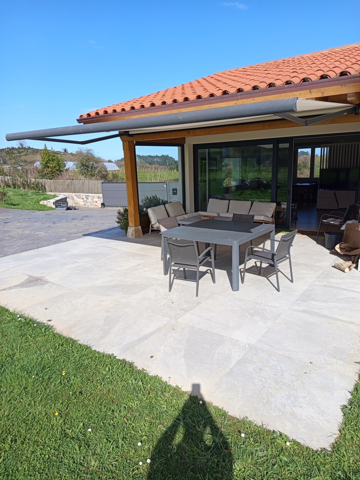An outdoor dining area is presented, featuring a rectangular table surrounded by six gray chairs. The space is shaded by a roof overhang, with a view of green grass and a clear blue sky visible in the background.