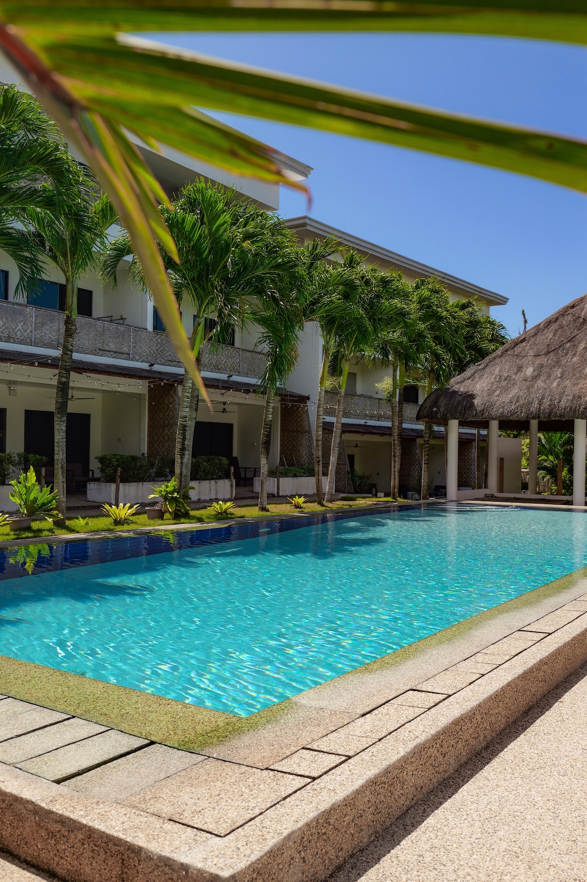 A clear swimming pool reflects the bright blue sky, surrounded by lush greenery and tropical palm trees. The area includes a thatched-roof structure nearby, providing shaded seating. The pool's edge is crafted with a smooth stone walkway, enhancing the inviting outdoor space.