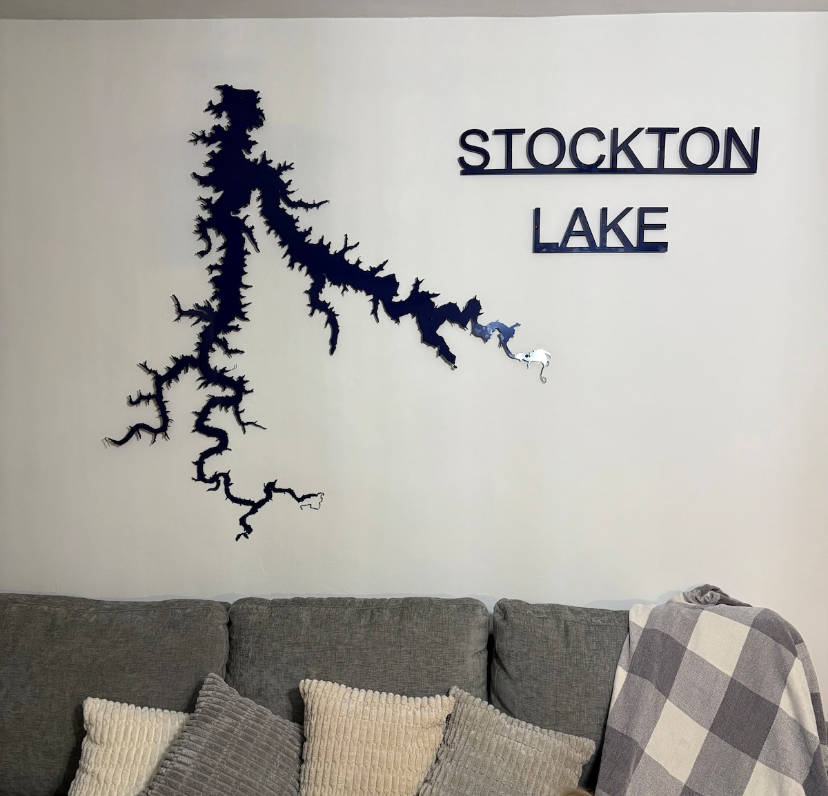 A wall displays a silhouette map of Stockton Lake, crafted in a deep blue color. The word 'STOCKTON LAKE' is prominently featured above the design. Below, a grey, textured sofa is adorned with various cushions and a cozy plaid throw for added comfort.