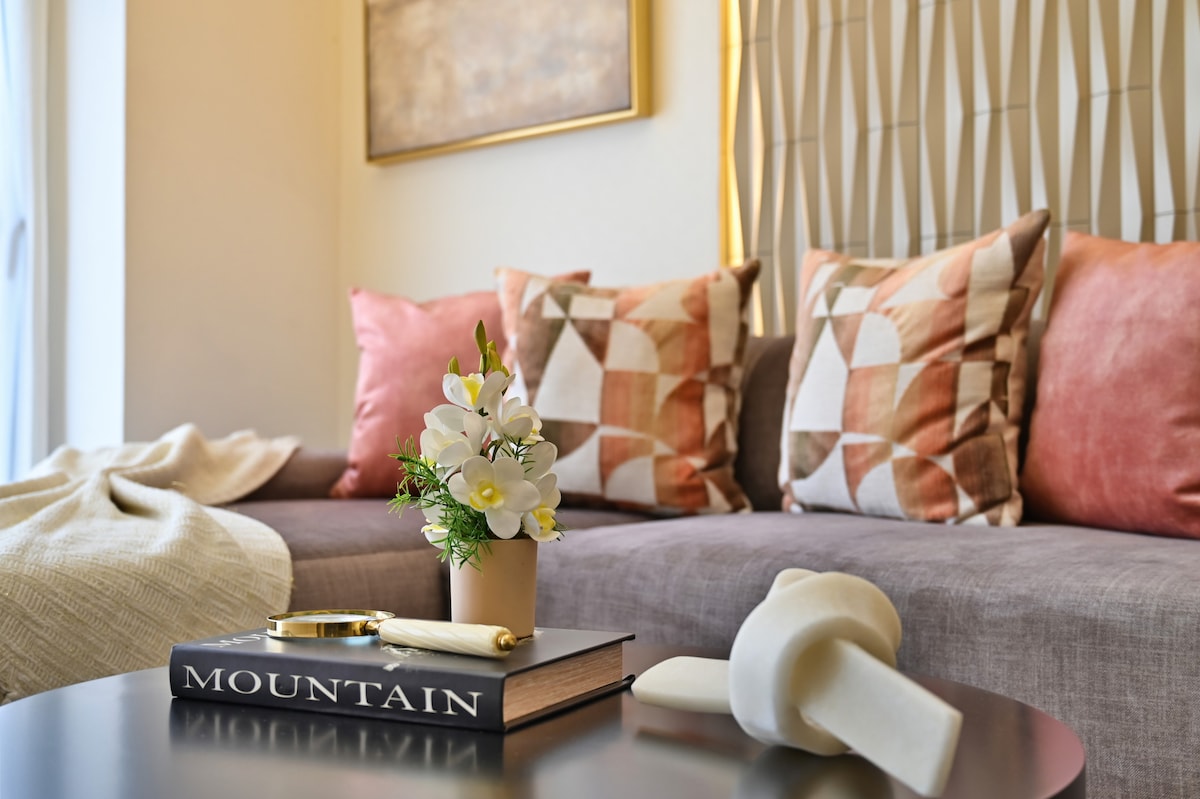 A comfortable seating area is highlighted by a neutral-toned sofa adorned with decorative pillows in shades of pink and brown. A small vase with white flowers sits atop a dark coffee table, accompanied by a book titled 'MOUNTAIN' and a decorative piece.