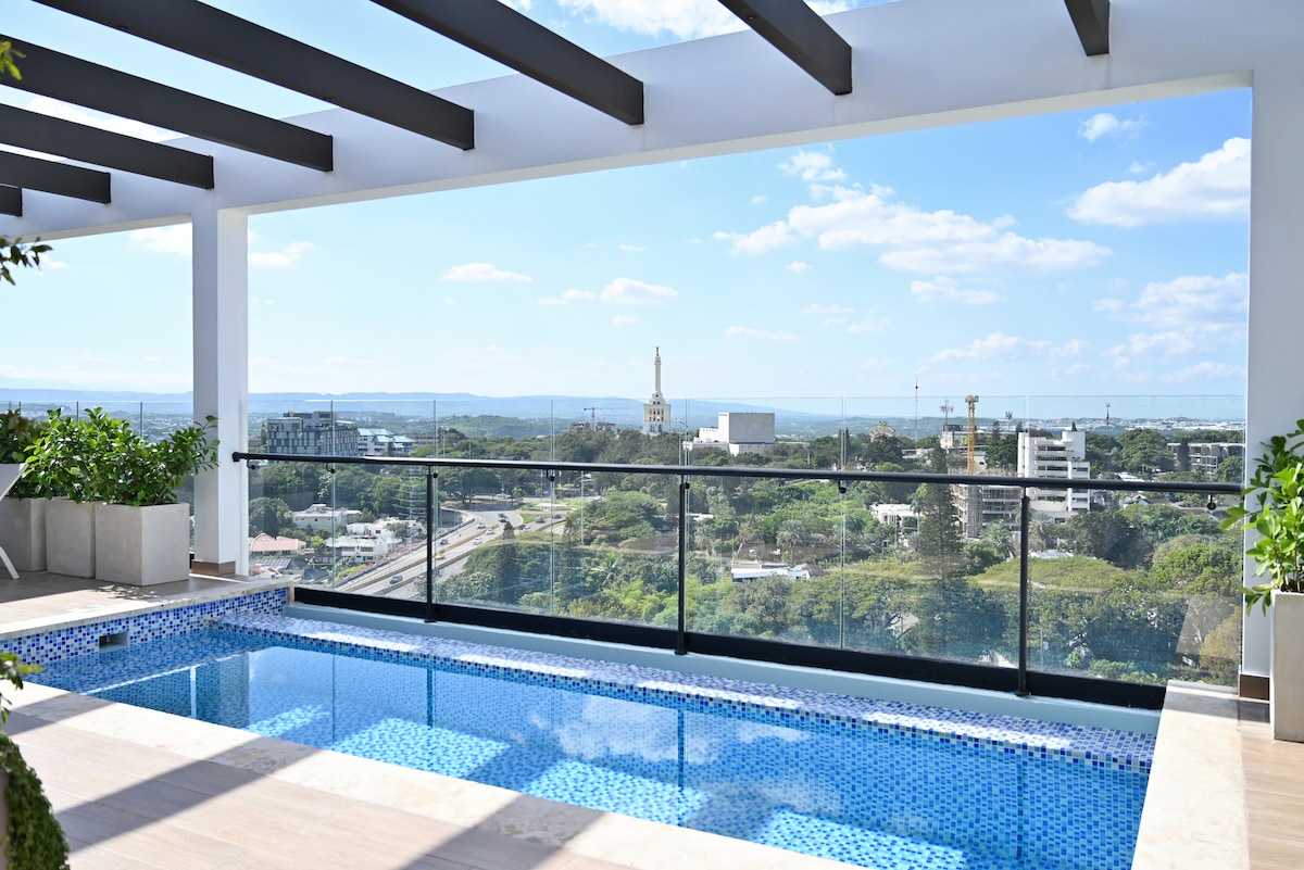 A spacious outdoor terrace is highlighted by a sleek pool, surrounded by vibrant greenery. The area features modern architecture with a glass railing that provides unobstructed views of the skyline and surrounding landscape, bathed in natural light.
