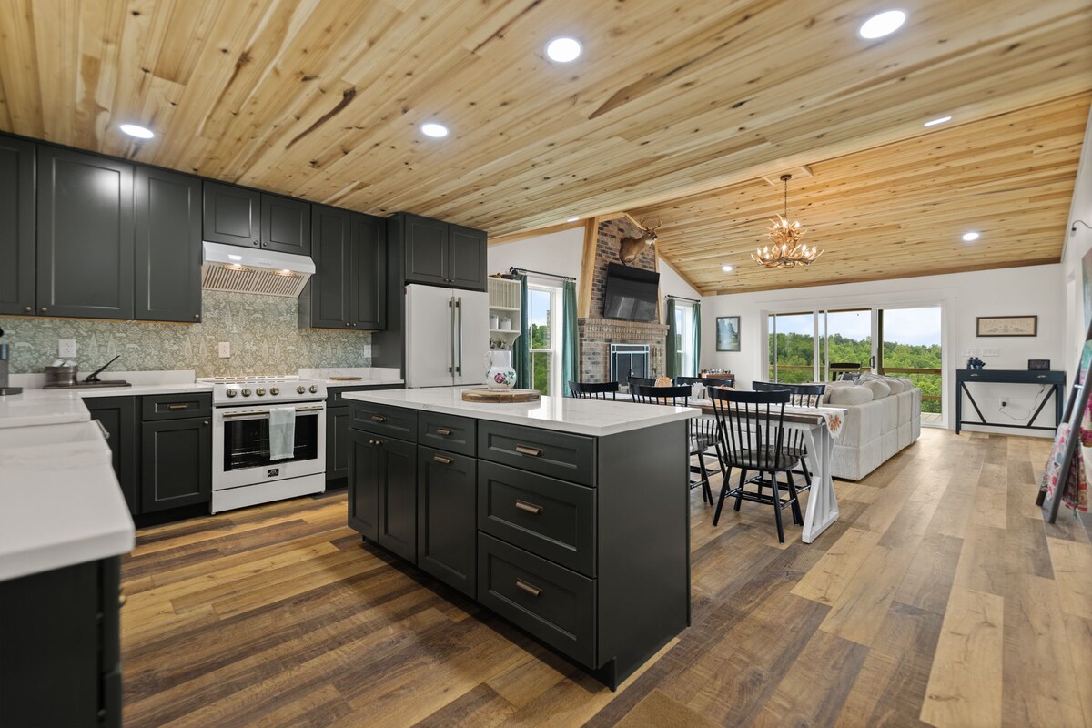 A spacious kitchen is featured with dark cabinetry and white countertops, complemented by a decorative backsplash. Modern appliances, including a stove and refrigerator, are visible. Adjoining areas reveal a cozy living space with large windows showcasing the natural landscape and wood accents throughout the ceiling.
