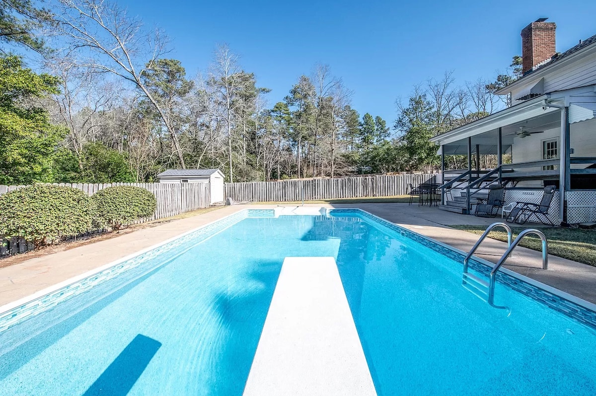 A spacious swimming pool is visible, surrounded by lightly landscaped areas with greenery and a wooden fence in the background. An adjacent covered patio area offers seating options, while a diving board is centered above the calm blue water.