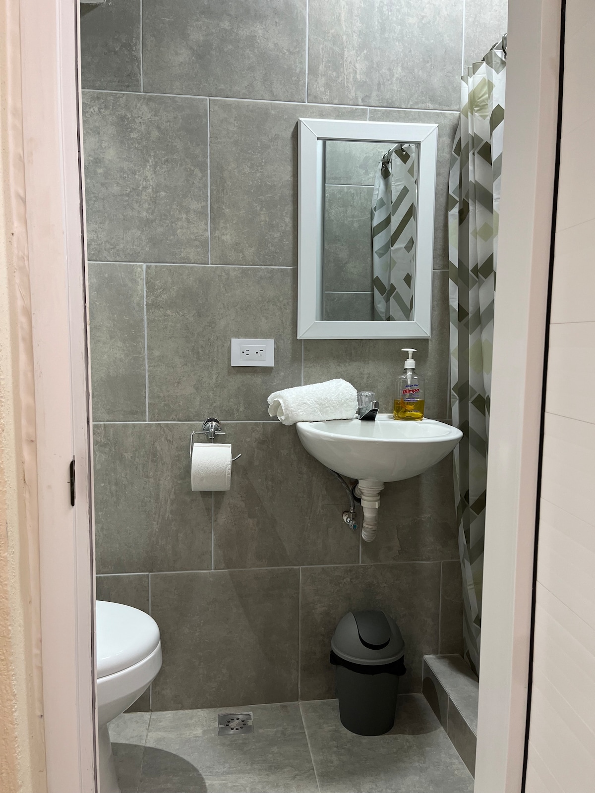 The compact bathroom is designed with neutral grey tiles, featuring a shower area with a patterned curtain. A small sink with modern fixtures is positioned next to a toilet. A mirror above the sink reflects the space, and essential toiletries are neatly arranged.